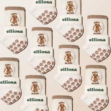 Multiple pairs of white and brown socks with 'elliossa' branding and brown dog illustrations embroidered, with brown paw print grips on the sole over a beige background.