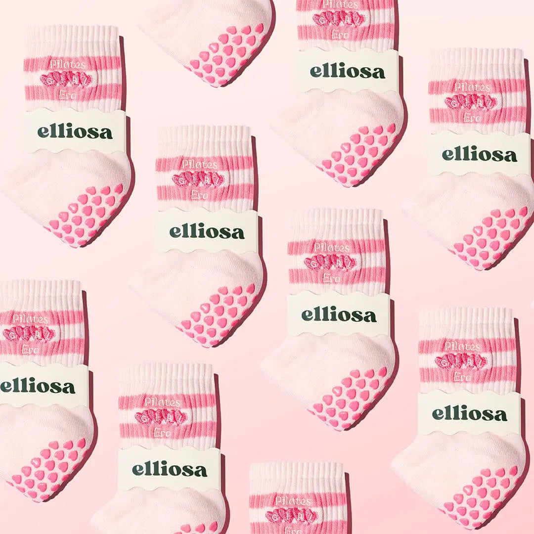 Pattern of pink and white socks with 'elliosa' branding in the packaging on a pink background. The socks have pink heart shaped grips on the soles and the words 'pilates girl era' embroidered with hearts on the ankle.
