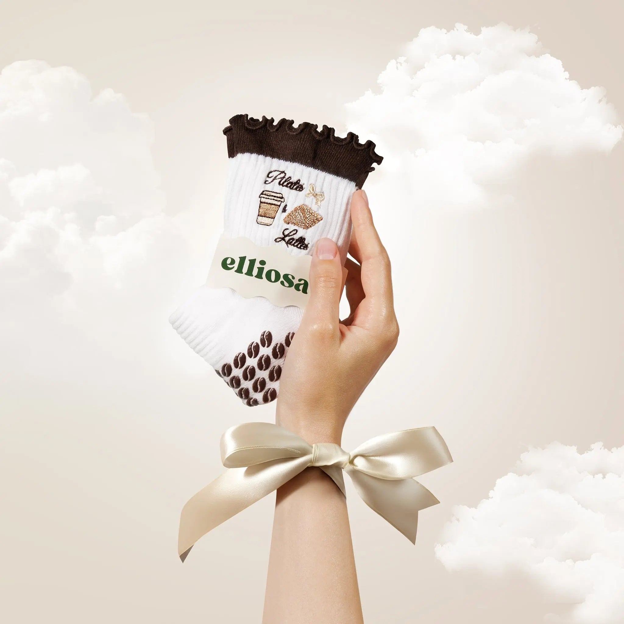 Hand holding white and brown patterned socks with 'elliosa' branding against a soft, cloud-like background.