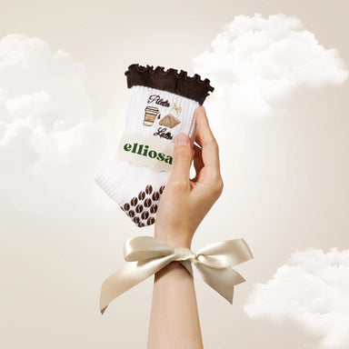 Hand holding white and brown patterned socks with 'elliosa' branding against a soft, cloud-like background.