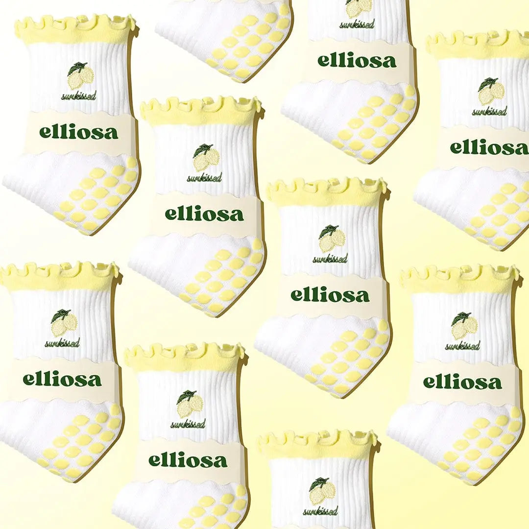 White socks with yellow frills and lemons and the word 'sunkissed' in green, with 'elliosa' branding against a light yellow background. There are yellow lemon shaped grips on the sole.