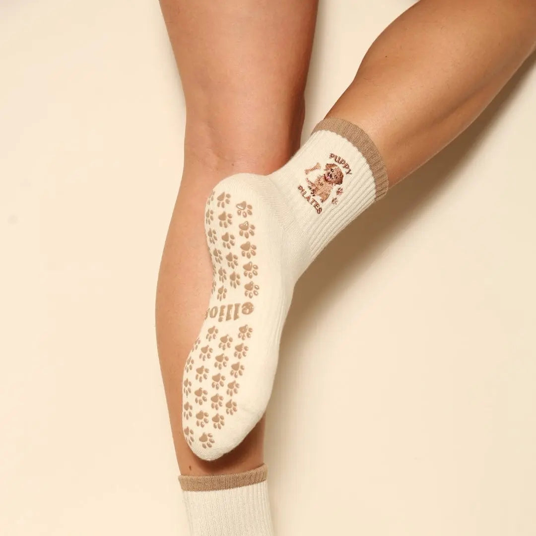 White socks with brown paw print grips on the sole and 'Puppy Pilates' text with an image of a puppy embroidered.