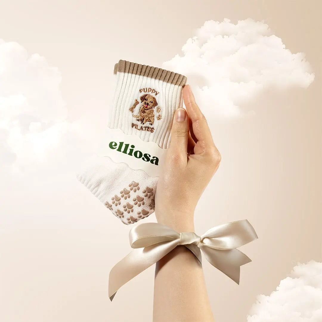 Hand holding white grip socks with a brown stipe at the top and a brown puppy embroidered with the words "puppy pilates" and brown paw print shaped grips on the sole, with 'elliosa' branding on the packaging.