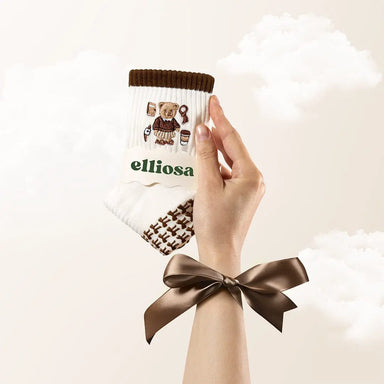 Hand holding white grip socks with a brown stripe at the top and brown bow shaped grips on the soles, and an image of a bear and coffee embroidered on the ankle.
