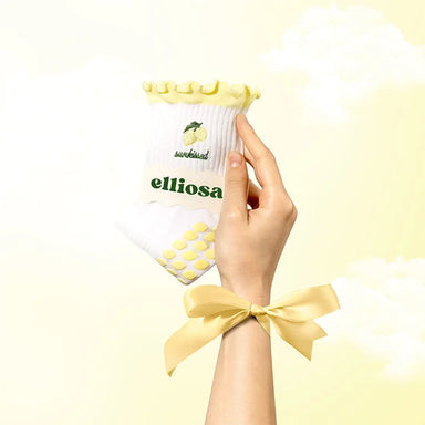 Hand holding a white product sock with yellow lemons and with the word 'sunkissed' with 'elliosa' branding against a light yellow background. There are yellow lemon shaped grips on the sole.