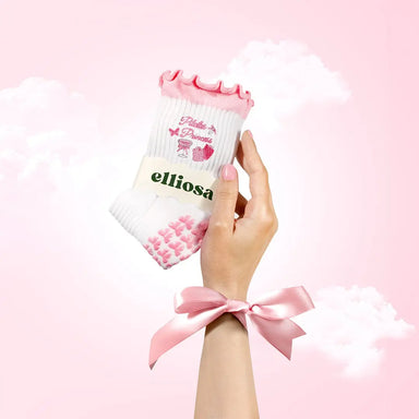 Hand holding a pair of white socks with pink accents and 'elliosa' branding against a pink background.