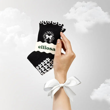 Hand holding a black and white patterned sock with the words "pilates social club" embroidered around a bow, in packaging with 'elliosa' branding against a light gray background.