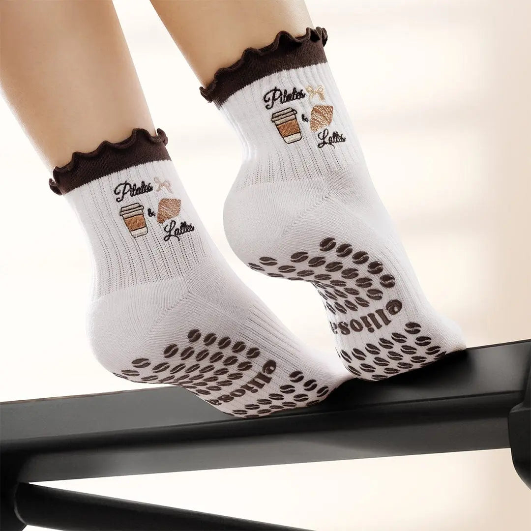 White socks with brown ruffles and an image of coffee and a croissant with the words "pilates & lattes" embroidered on the ankle, and coffee bean shaped grips on the soles. 