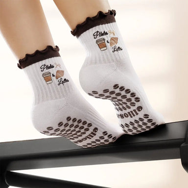White socks with brown ruffles and an image of coffee and a croissant with the words "pilates & lattes" embroidered on the ankle, and coffee bean shaped grips on the soles. 