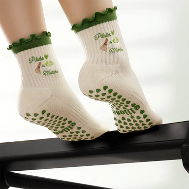 White socks with green heart shaped grips on the soles, with the text 'pilates & matcha' embroidered in green with images of matcha on the ankles.