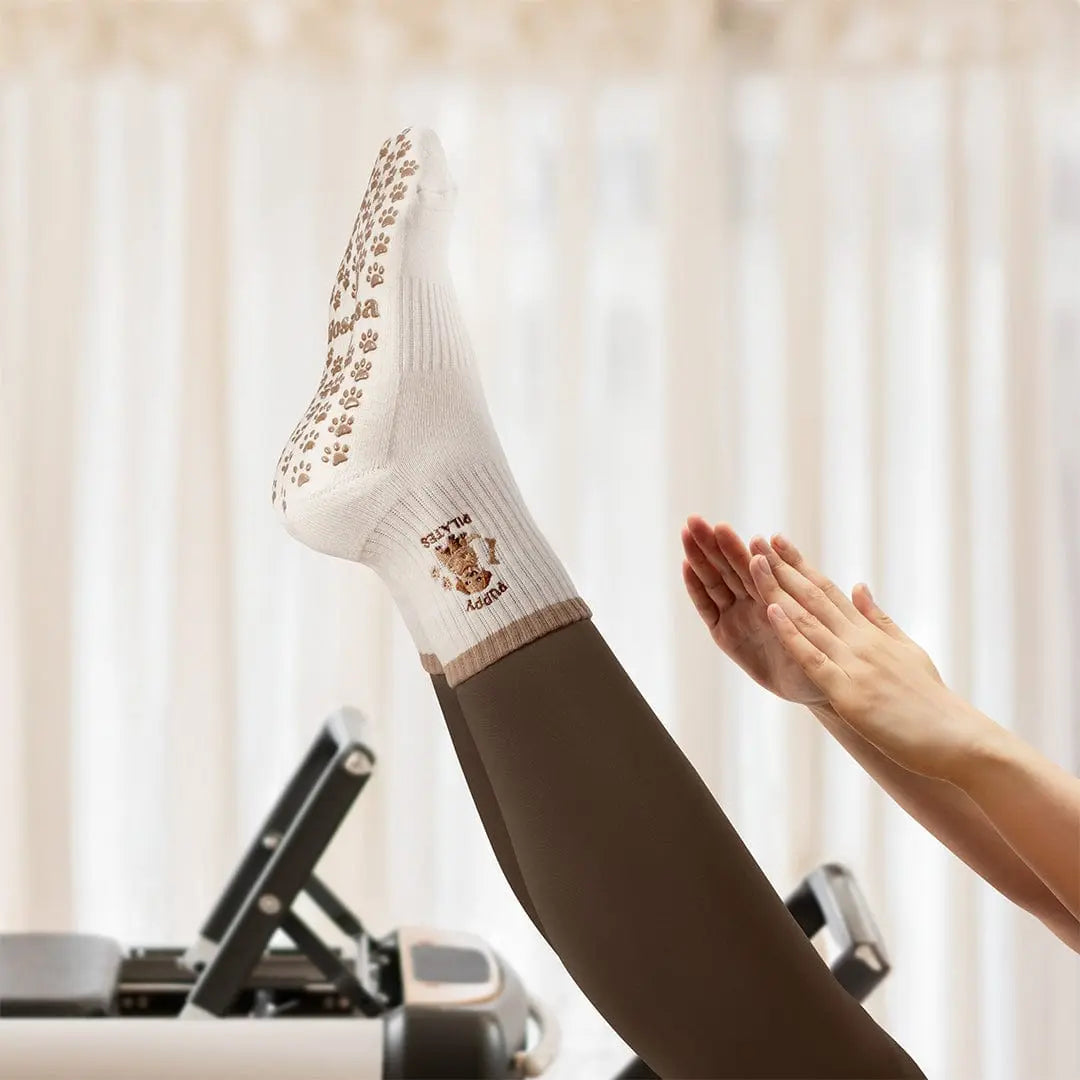 Person on a reformer pilates bed while wearing brown leggings and white grip socks with a puppy image and 'puppy pilates' embroidered.