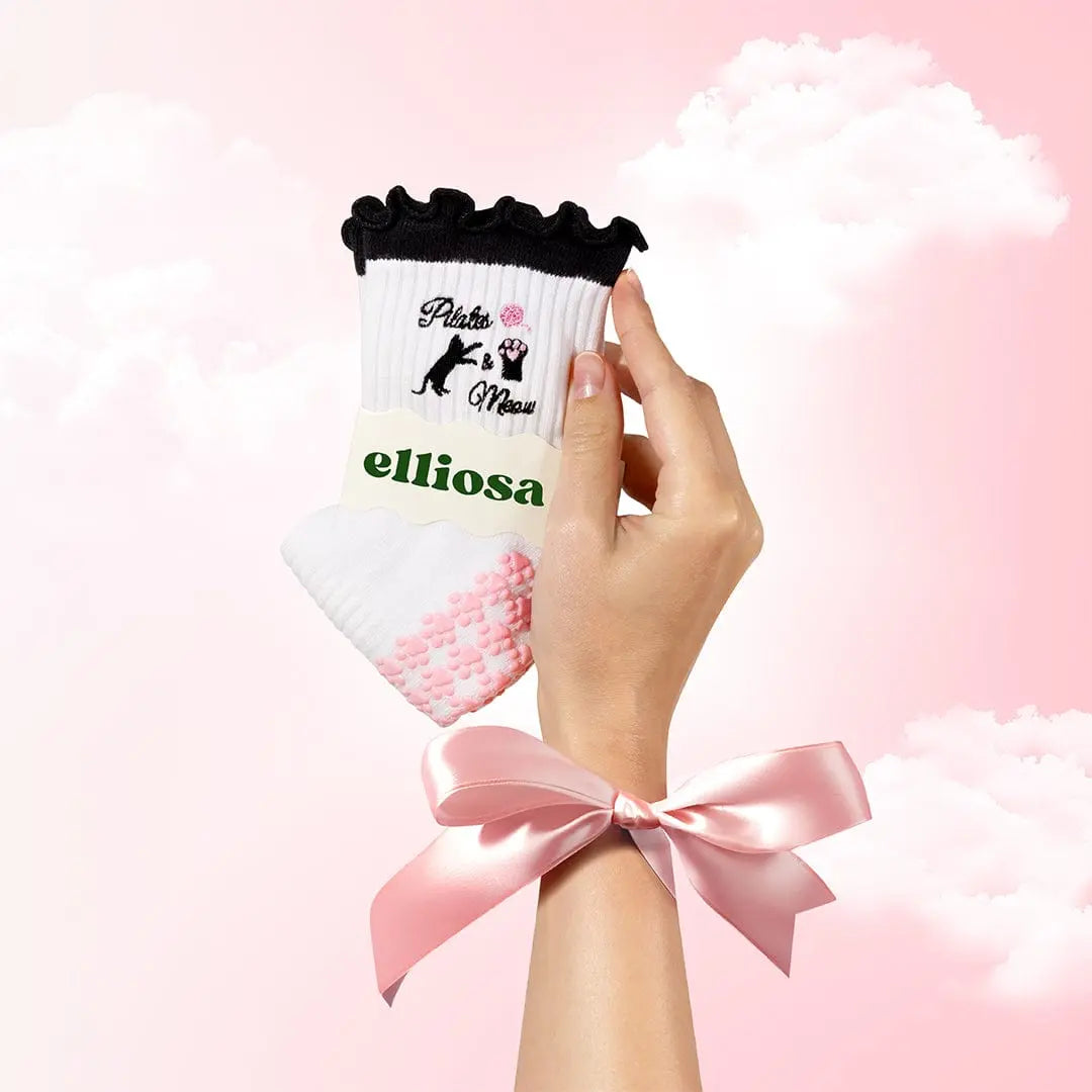 Hand holding a white sock with black ruffle top with pink paw shaped grips on the soles, and black "pilates and meow" text with images of a cat. 