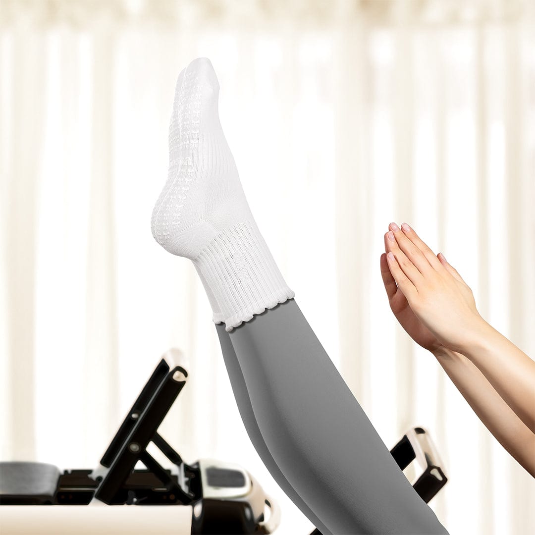 Person using a Pilates reformer machine with one leg raised wearing a white sock and gray leggings.
