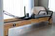 Angled view of the Core Collab Eco Reformer showing Beechwood frame with charcoal upholstery in a room with concrete floor and sheer curtains.