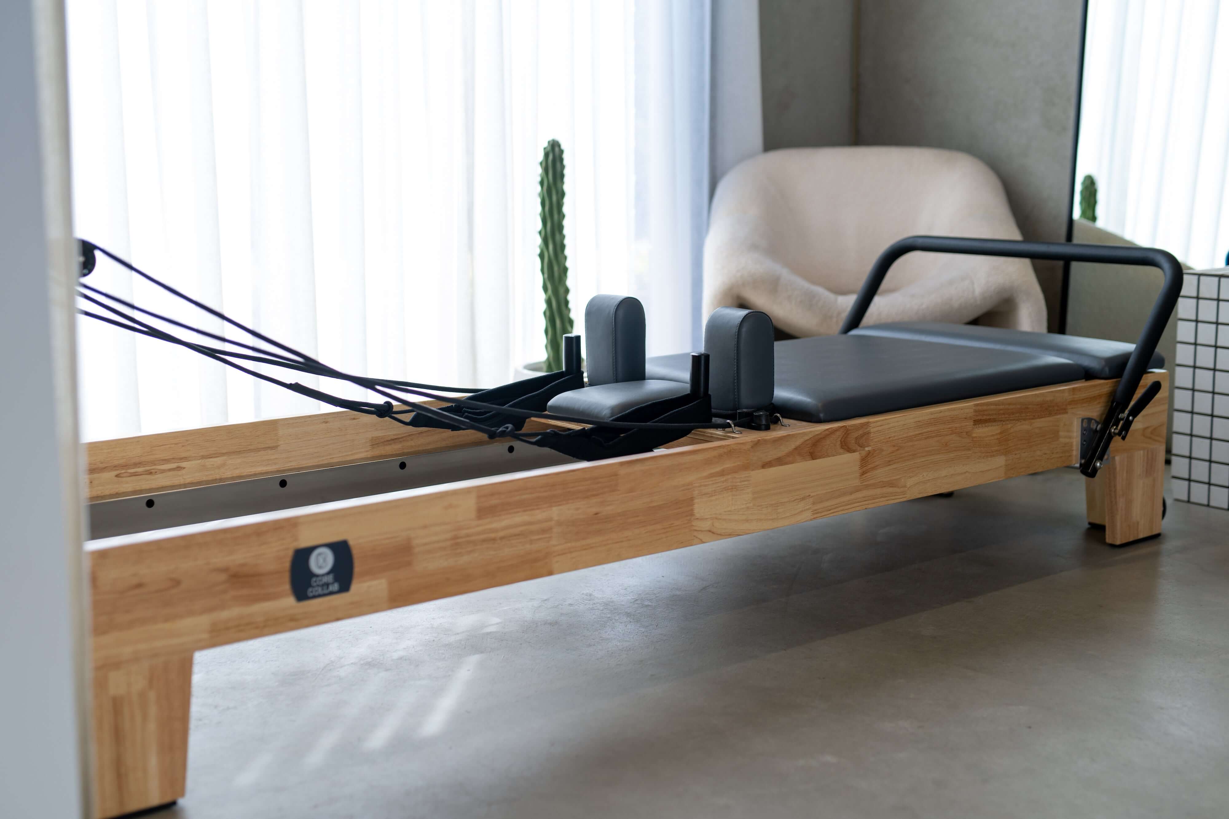 Angled view of the Core Collab Eco Reformer showing Beechwood frame with charcoal upholstery in a room with concrete floor and sheer curtains.