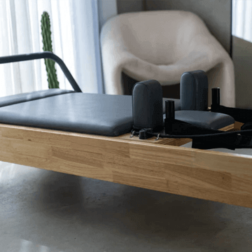 Close up of the Core Collab Eco Warrior Pilates Reformer showing a Beechwood frame and charcoal upholstery in a room with a cream coloured chair.