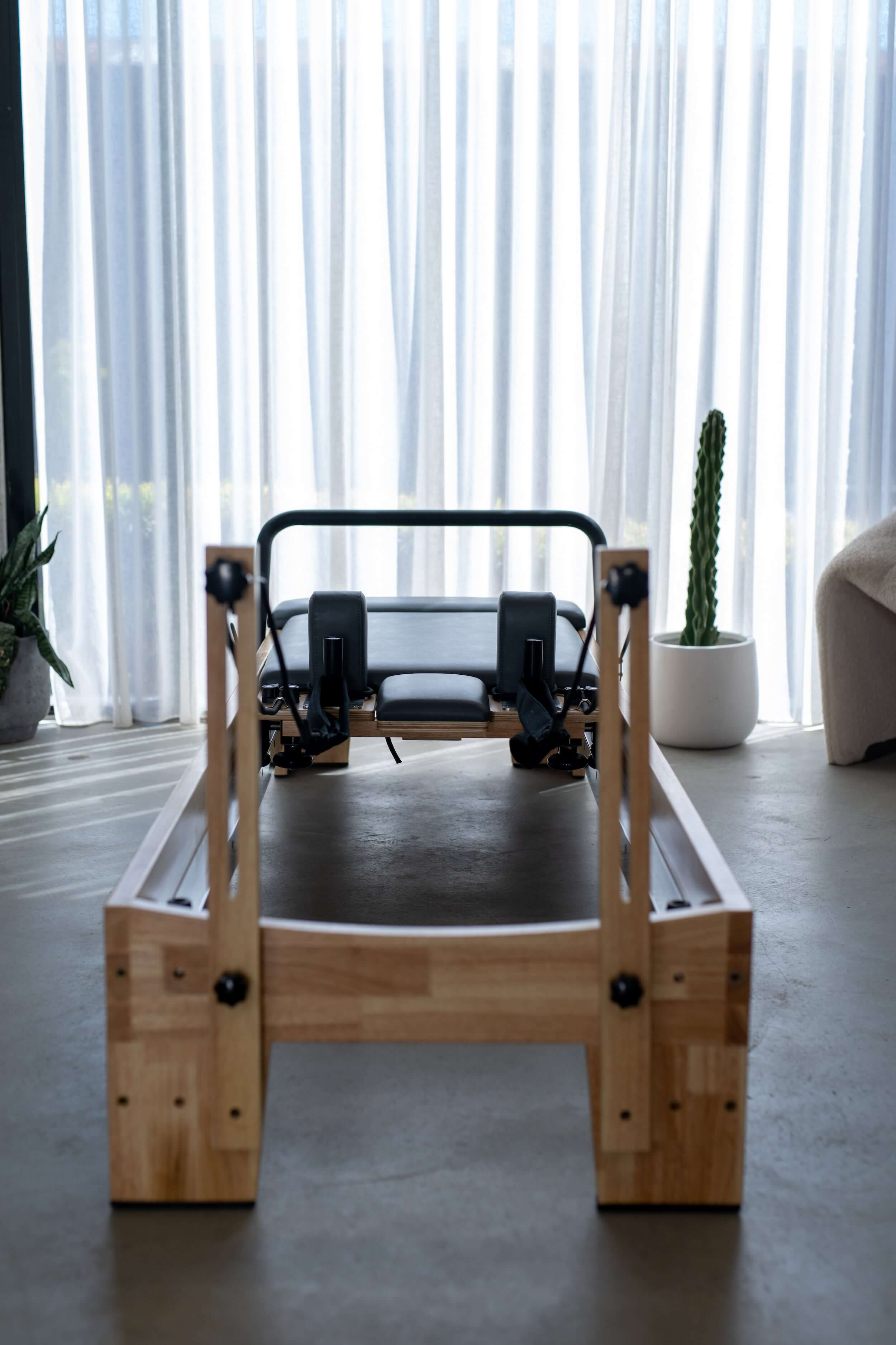 Core Collab Eco Pilates reformer view from the end showing Beechwood and black upholstery, in a room with concrete floor and sheer curtains.