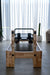 Core Collab Eco Pilates reformer view from the end showing Beechwood and black upholstery, in a room with concrete floor and sheer curtains.