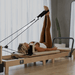 Woman using a Core Collab foldable eco Pilates reformer in beige with feet in straps.