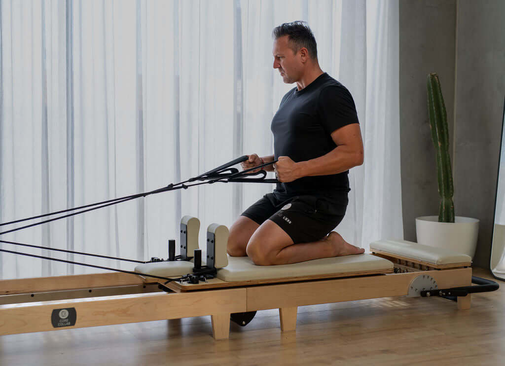Man using a Core Collab Foldable Eco Pilates reformer in beige, kneeling with hands in straps.