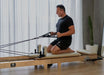 Man using a Core Collab Foldable Eco Pilates reformer in beige, kneeling with hands in straps.