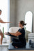 Woman sitting on a Core Collab Queen Pilates Reformer Tower with legs crossed and hands in straps.