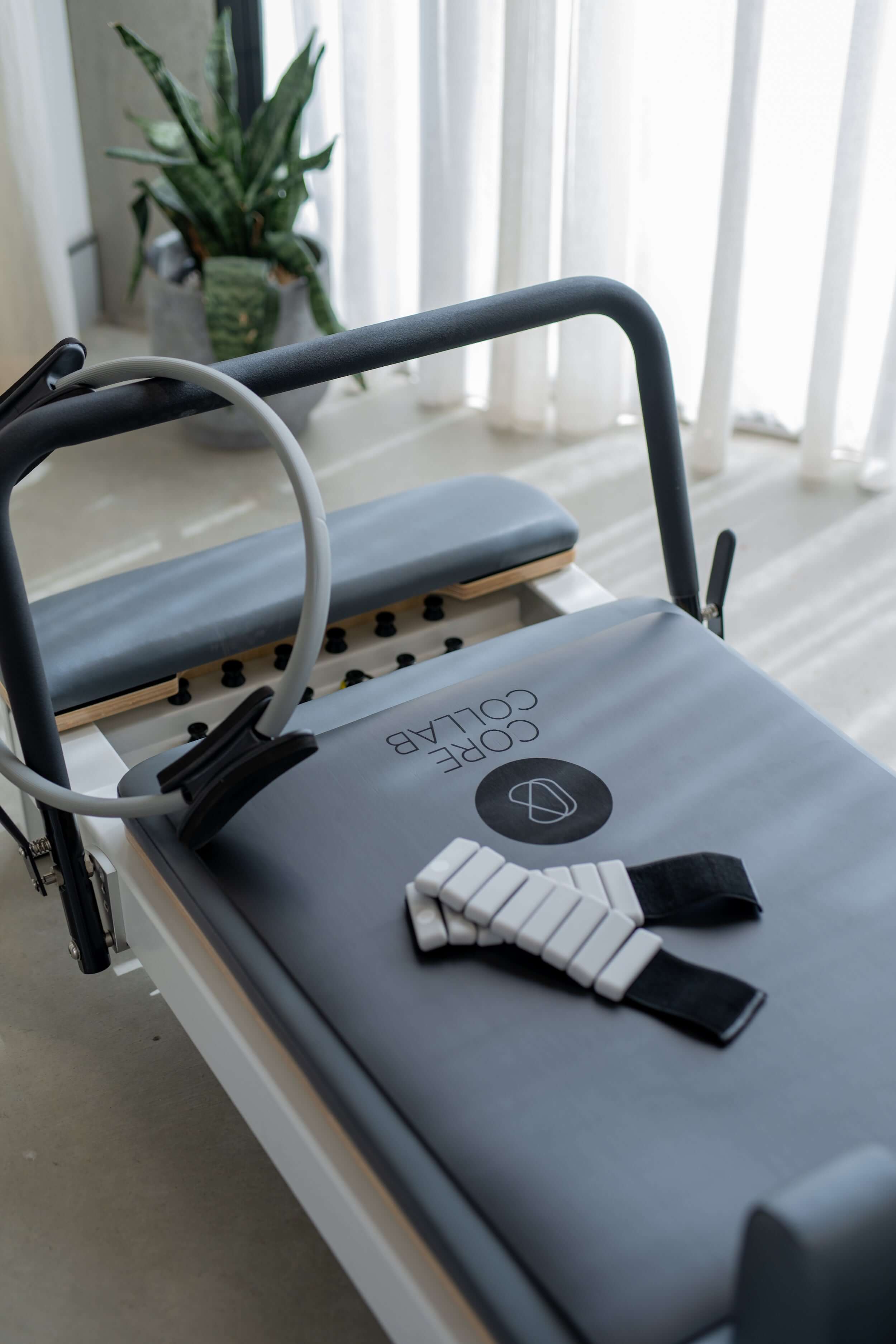 The Core Collab Queen Reformer with tower shown in white with grey upholstery at the footbar end with a Pilates magic circle and ankle weights.
