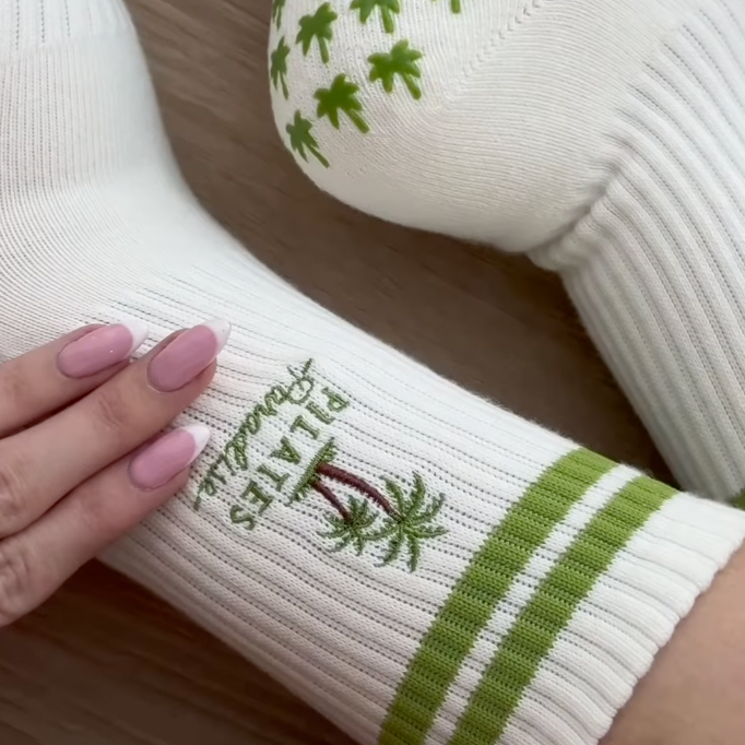 Close up of Pilates paradise socks on feet, showing the embroidered palm tree and Pilates Paradise text, and palm shaped grips on the soles.