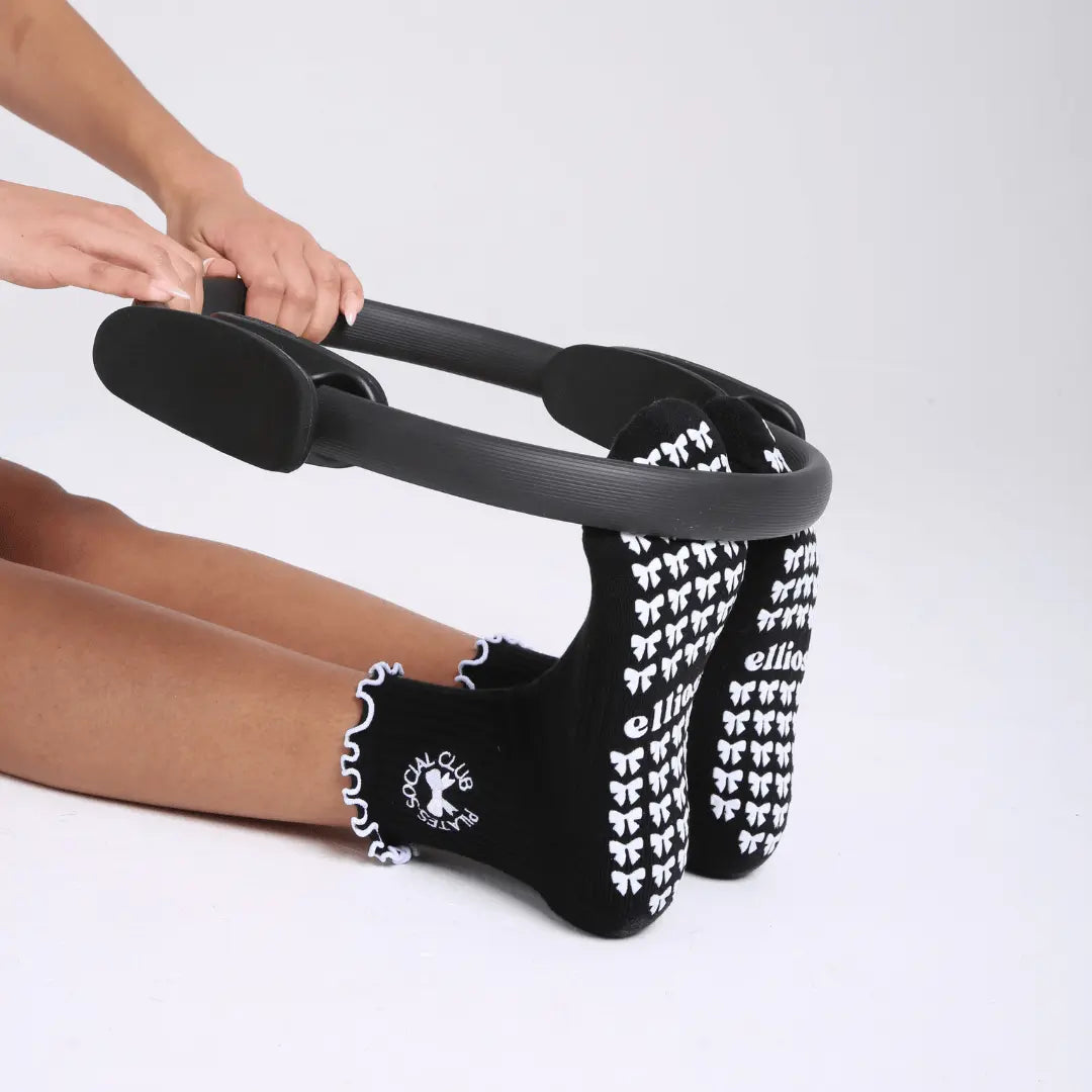 Person using a black pilates ring with black Elliosa dual direction grip socks with white bow shaped grips on the soles, with white ruffles and 'pilates social club' embroidered around a bow on the ankle. 