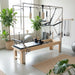 Full trapeze reformer in bright studio setting with greenery and grey Pilates accessories.
