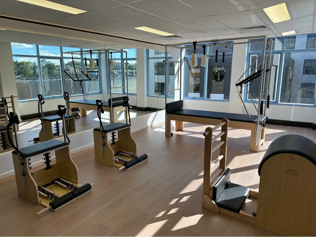 Wooden Cadillac Table with black upholstery in a studio with large windows surrounded by other pilates equipment.