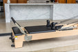 Close up of light wood coloured pilates reformer with black upholstery, showing foot bar and shoulder pads.