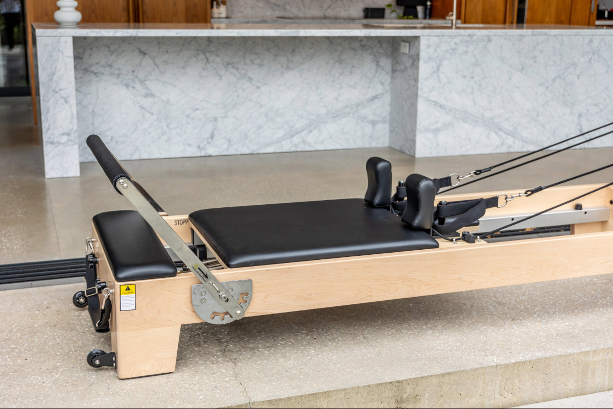 Close up of light wood coloured pilates reformer with black upholstery, showing foot bar and shoulder pads.