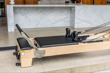 Close up of light wood coloured pilates reformer with black upholstery, showing foot bar and shoulder pads.