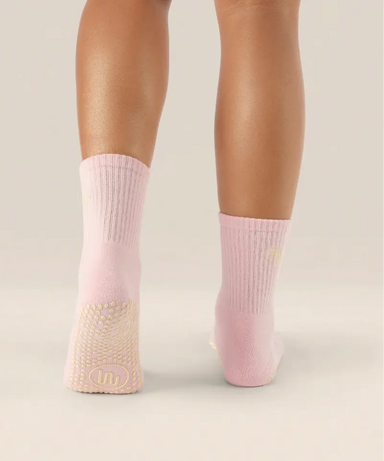 Rear view of soft pink coloured crew grip socks above the ankle with "pilates club" in yellow text on the ankle and yellow grips on the sole.
