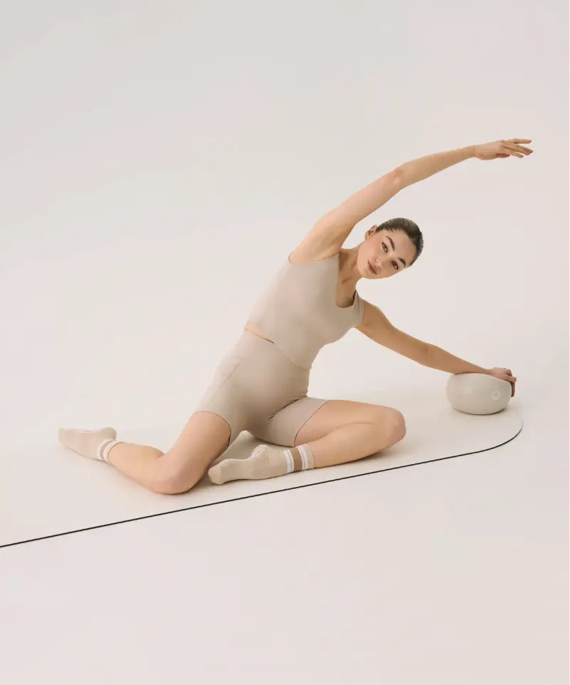 Woman doing pilates on a mat wearing beige outfit with matching beige socks with white stripes.