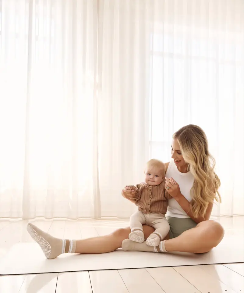 Woman sitting holding a baby on a pilates mat wearing beige grip socks with white strips and white sole grips.