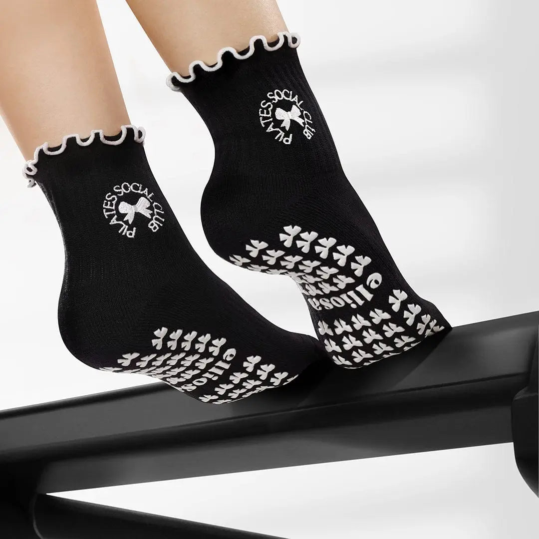 Black Elliosa dual direction grip socks in Pilates Social Club with white bow-shaped grips on the sole and 'pilates social club' embroidered around a bow.