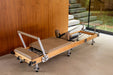 Foldable wooden reformer pilates machine with light brown upholstery, wooden frame with steel elements and black pulleys