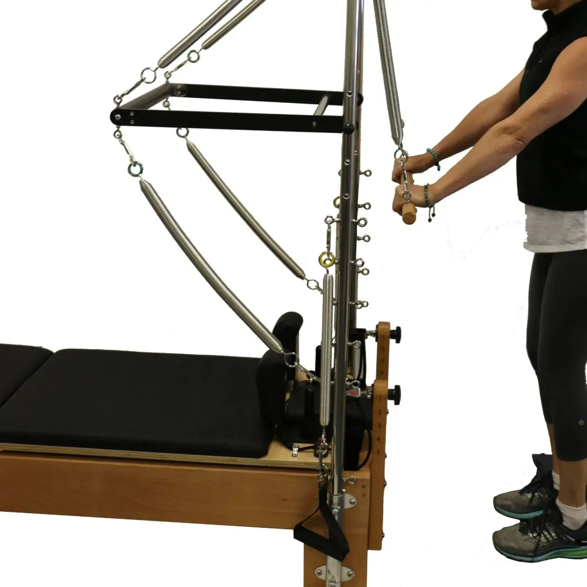 Close up of wooden pilates reformer table with black upholstery. A person is using the trapeze attachment.