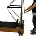 Close up of wooden pilates reformer table with black upholstery. A person is using the trapeze attachment.