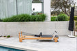 Light wood coloured pilates reformer with black upholstery on a patio with a pool and garden in the background
