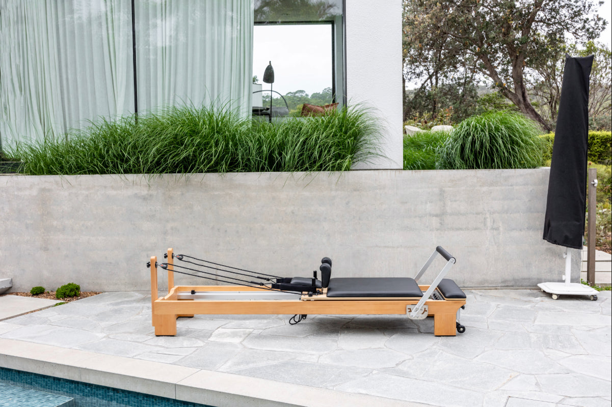 Light wood coloured pilates reformer with black upholstery on a patio with a pool and garden in the background