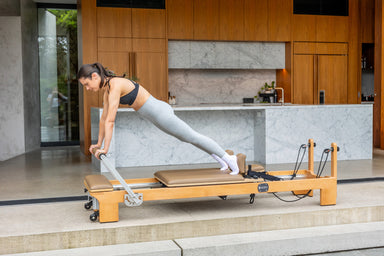 Woman exercising on a light brown coloured Pilates reformer in a modern kitchen.