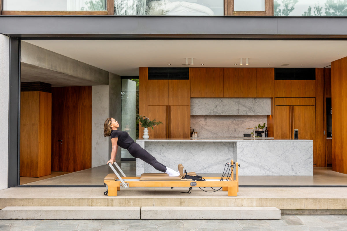 Woman using a wooden pilates reformer machine with light brown upholstery in a modern home setting