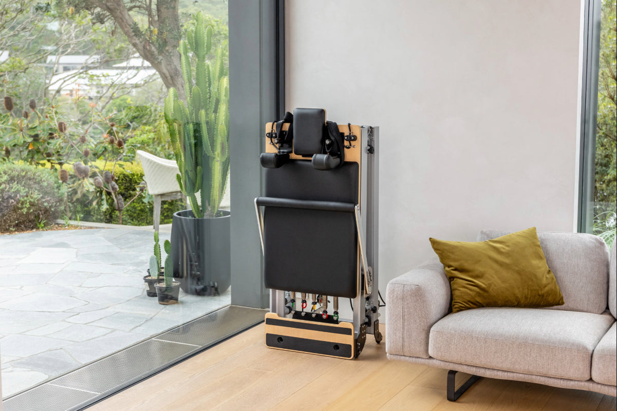 Folded reformer pilates machine in corner of modern living room. The reformer is made of metal, light coloured wood and has black upholstery. There is greenery outside.