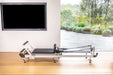 Metal and black pilates reformer machine in a room with a television and window view of greenery.