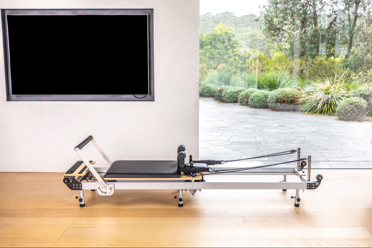 Metal and black pilates reformer machine in a room with a television and window view of greenery.