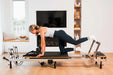 Woman exercising on a metal and black Pilates reformer machine in a home setting.