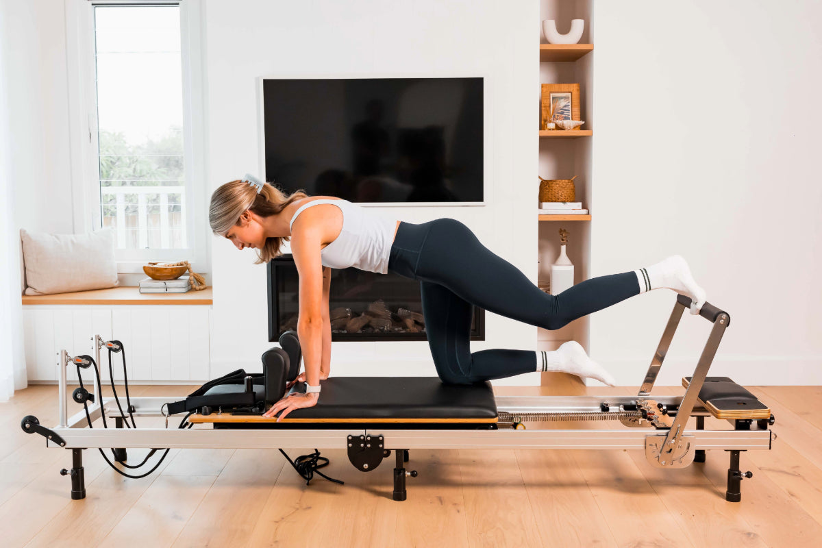 Woman exercising on a metal and black Pilates reformer machine in a home setting.